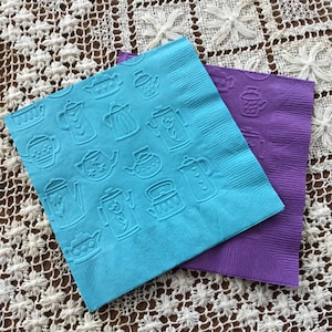 May include: Two paper napkins, one blue and one purple. The blue napkin has a pattern of embossed teapots and teacups. The purple napkin is plain.