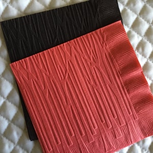 May include: Two decorative paper napkins. One is coral-colored, and the other is black. Both napkins have a textured, embossed pattern of vertical lines and curved shapes. The napkins are stacked on top of each other.