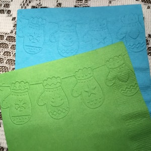 May include: Two green and blue paper napkins with a white embossed design of mittens hanging on a string. The mittens have different designs, including snowflakes, stars, and hearts.