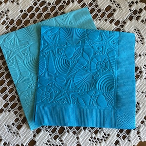 May include: Two blue paper napkins with an embossed design of seashells and starfish. The napkins are folded in half and are on a white crocheted surface.