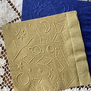 May include: A gold-colored paper napkin with an embossed design of constellations, stars, planets, and moons. The napkin is folded in half and has a textured surface.