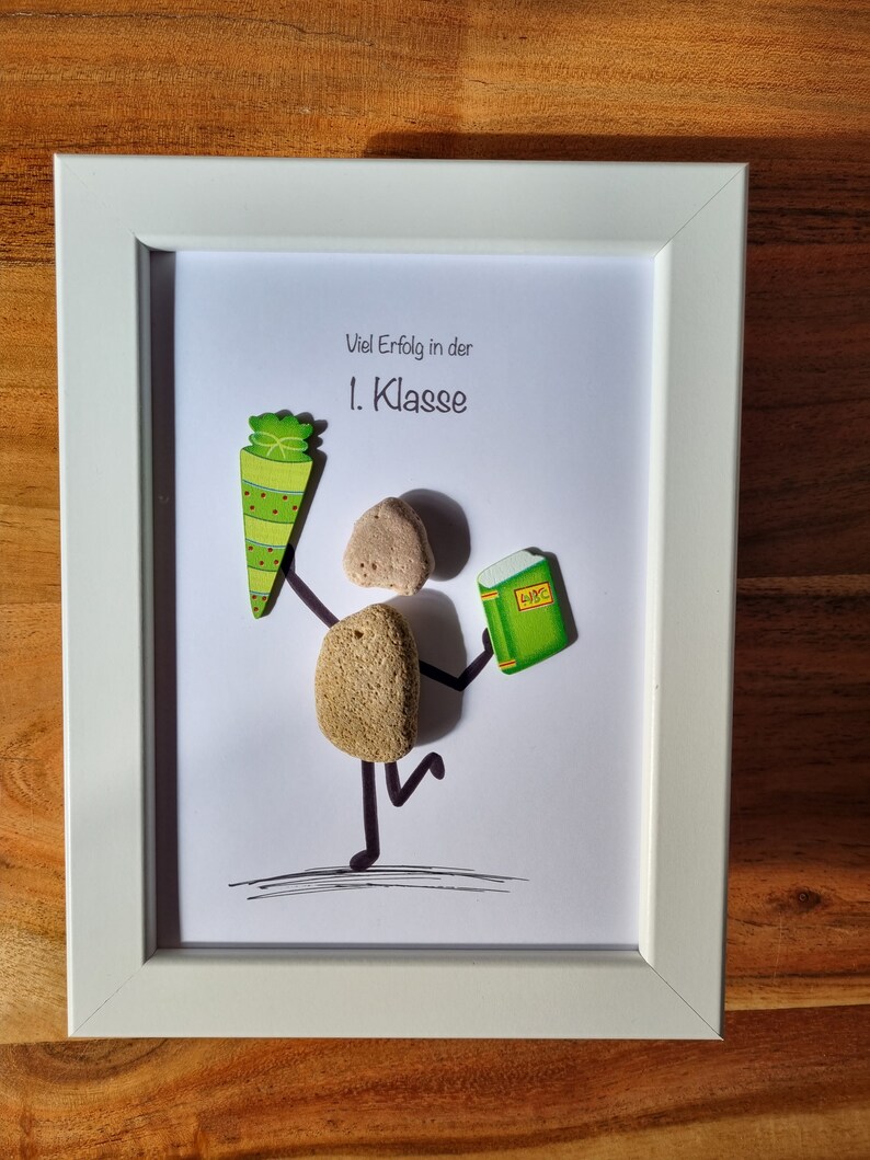 May include: A white frame with a white mat and a white background. The background has a green and red school cone, a brown rock with a black line drawn on it, and a green book with the word "Buch" written on it. The text "Viel Erfolg in der 1. Klasse" is written in black on the white background.