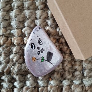 以下が含まれることがあります： 紫色の背景に、黒い目、黒い鼻、黒と緑の花が描かれた手描きの石。石は茶色の織り地の上に置かれています。