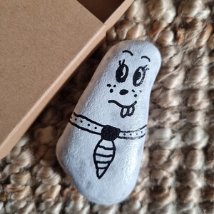 以下が含まれることがあります： 手描きの白い石で、漫画の顔、黒い目、舌が出ています。黒いネクタイと襟が付いています。石は、小さな開いた段ボール箱の隣にあります。