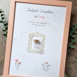 Könnte beinhalten: Ein rosa Rahmen mit weißem Hintergrund und einem Zertifikat für die Zahnfee. Das Zertifikat lautet "Zahnfee Zertifikat" und "für Emily". Der Text lautet außerdem "Dein erster Zahn ausgefallen am: 20. September 2023". Es gibt einen kleinen weißen Rahmen mit einem Glasgefäß darin. Das Gefäß hat einen Korkverschluss. Auf der linken Seite des Rahmens befindet sich eine kleine rosa Blume.