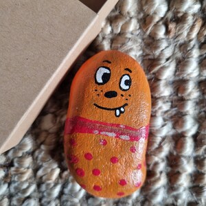 以下が含まれることがあります： 手描きで顔を描いた石。オレンジ色の石に赤い水玉模様と、中央に赤と白のストライプが描かれている。顔は白い目で、黒い鼻と白い口に2本の歯がある。