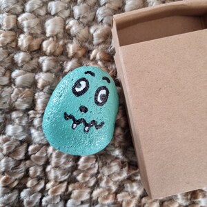 以下が含まれることがあります： 漫画の顔を描いたターコイズグリーンに塗られた石。石には大きな目が2つ、小さな歯のついた口、小さな鼻があります。石は茶色の織り地の上に置かれています。