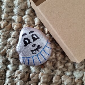 以下が含まれることがあります： 銀色に塗られた石に、漫画の顔が描かれている。顔は、大きな目が2つ、小さな鼻、歯の生えた大きな口をしている。石は茶色の織り方の敷物の上に置かれている。