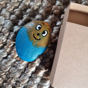 以下が含まれることがあります： 青と黄色の顔を描いた手描きの石。石は茶色の織り絨毯の上に、小さな茶色の箱の隣に置かれています。