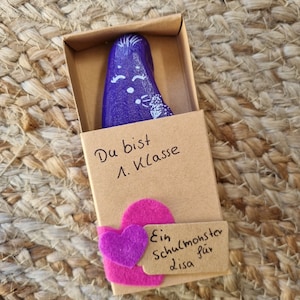 以下が含まれることがあります： 茶色のマッチ箱の中に、顔とハートが描かれた小さな紫色の石が入っています。箱にはピンクのハートのステッカーと、「Du bist 1. Klasse Ein Schulmonster für Lisa」と書かれた手書きのメモが付いています。