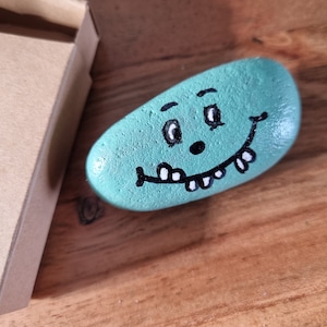 以下が含まれることがあります： ターコイズブルーに塗られた石に、笑顔の顔が描かれています。顔には、目、鼻、歯の生えた口があります。