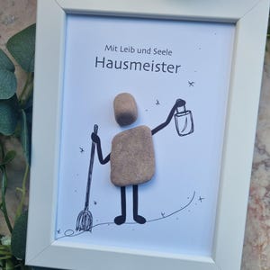 Könnte beinhalten: Gerahmtes Kunstwerk mit einer Kieselstein-Figur eines Hausmeisters, der einen Besen und eine Laterne hält. Der Text "Mit Leib und Seele Hausmeister" ist über der Figur zu sehen. Der Rahmen ist weiß.