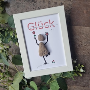 Puede incluir: Un marco blanco con una obra de arte que presenta una figura de piedra sosteniendo una rosa y un corazón. La palabra alemana "Glück" está escrita en la parte superior, con texto adicional debajo. Rodeado de vegetación.