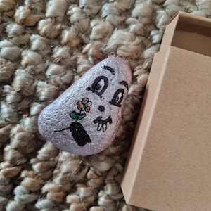 以下が含まれることがあります： 顔と花が描かれた石。石は薄い紫で、黒い絵の具で詳細が描かれている。石は茶色の織り地の上に置かれている。