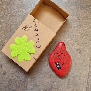 以下が含まれることがあります： 緑の四つ葉のクローバーと「Dein Glücksbringer」という文字が書かれた小さな茶色の箱。箱が開いており、隣には黒いテントウムシが描かれた赤い石が置かれている。