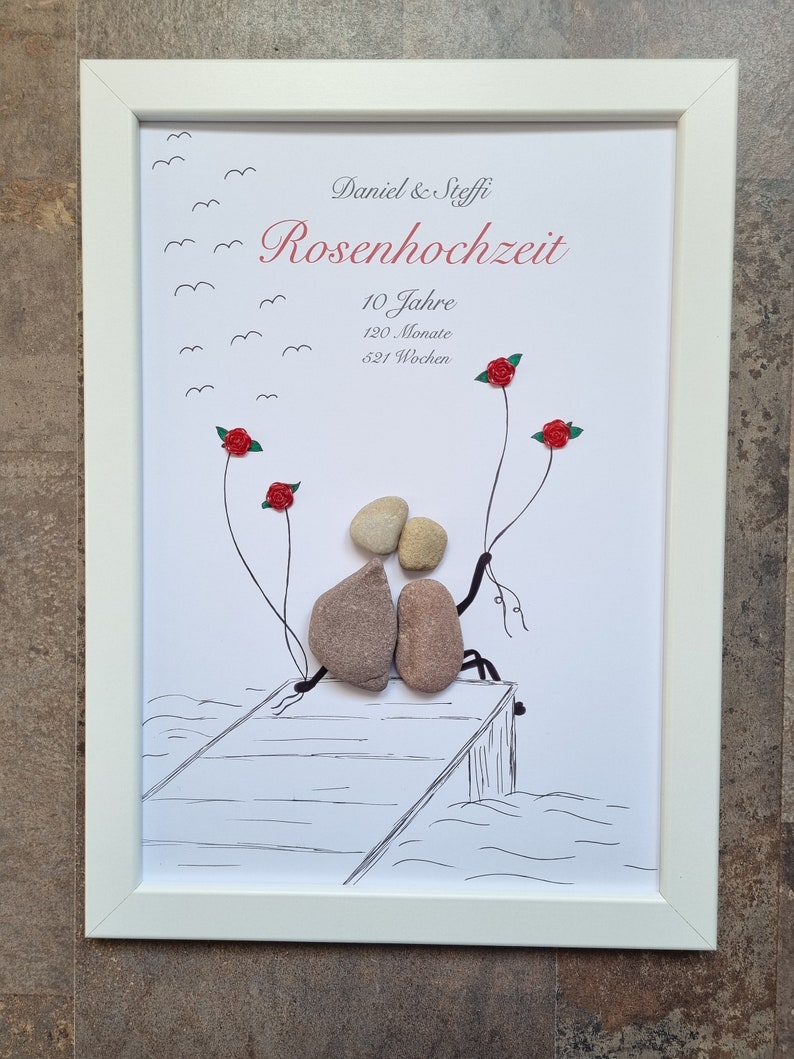 Puede incluir: Una imagen enmarcada en blanco con un dibujo en blanco y negro de dos personas sentadas en un muelle. Las personas est&aacute;n hechas de piedras y sostienen flores rojas. El texto en la imagen dice "Daniel & Steffi Rosenhochzeit 10 Jahre 120 Monate 621 Wochen."