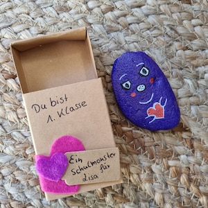 以下が含まれることがあります： 小さな茶色の箱に、顔と赤いハートが描かれた紫色の石が入っています。箱にはピンクのハートと、「Ein Schulmonster für Lisa」と書かれたタグが付いています。箱には「Du bist 1. Klasse」という文字も書かれています。