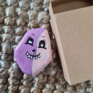以下が含まれることがあります： 紫とピンクの顔に黒い目と白い笑顔が描かれた石。石は茶色の織り目の表面に、小さな茶色の箱の隣に置かれている。
