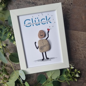 Pode incluir: Uma obra de arte emoldurada com uma borda branca. A obra apresenta uma figura de arte de seixos segurando um coração vermelho. O texto "Glück" e outro texto alemão estão acima da figura. A figura é feita de seixos e tem um rosto sorridente.