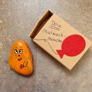 以下が含まれることがあります： かわいい顔と赤いハートを持ったオレンジ色の石が描かれています。石は、「Dein Mutmach-Monster」と書かれた小さなダンボール箱と赤いフェルトのハートの前にあります。