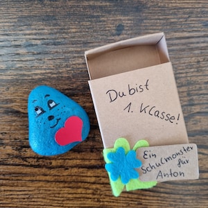 以下が含まれることがあります： 青い顔料で塗られた石に、笑顔と赤いハートが描かれている。石は小さな段ボール箱に入っていて、箱には「Du bist 1. Klasse!」という手書きのメッセージと、「Ein Schulmonster für Anton」と書かれた緑色のフェルトタグが付いている。