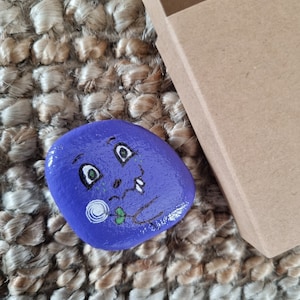 以下が含まれることがあります： 手描きの滑らかな紫色の石で、気まぐれな顔のデザインが施されています。顔には黒い目、白い渦巻き、小さな緑の葉があります。石は、小さな開いた茶色の段ボール箱の隣にあります。