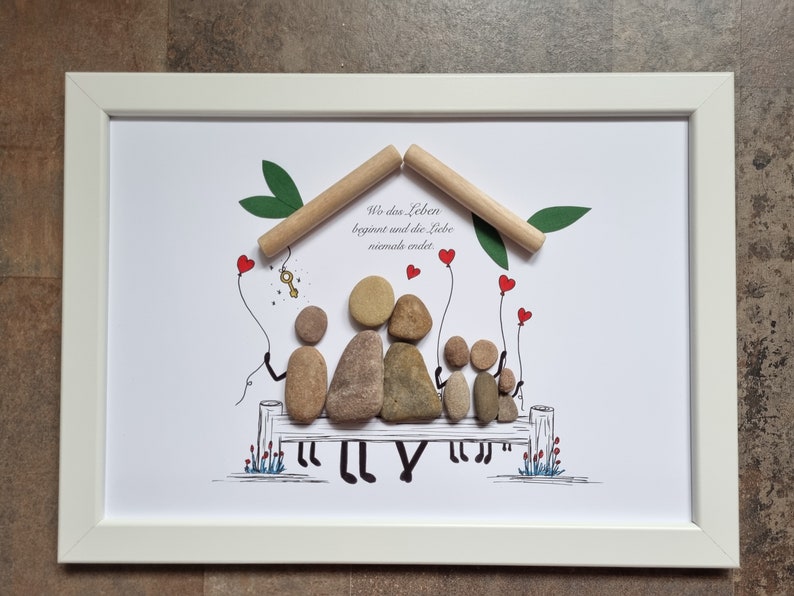 May include: A framed print with a hand-drawn illustration of a family sitting on a bench under a house made of sticks. The family is made of painted stones. The text "Wo das Leben beginnt und die Liebe niemals endet" is written on the print.