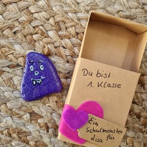 以下が含まれることがあります： 木製のマッチ箱の中に、紫色の絵の具で描かれた小さな石があり、漫画の顔が描かれている。マッチ箱にはピンクのフェルトのハートと、「Ein Schulmonster für Lisa」と書かれたタグが付いている。マッチ箱には、「Du bist 1. Klasse」という文字も書かれている。
