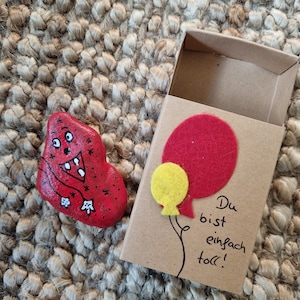 以下が含まれることがあります： 顔と星が描かれた小さな赤い石。石は、赤と黄色のフェルトの風船と「Du bist einfach toll!」という文字が書かれた小さな段ボール箱に入っています。