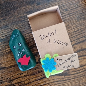 以下が含まれることがあります： 緑色の顔と赤いハートが描かれた小さな石。石は小さな段ボール箱に入っていて、箱には「Du bist 1. Klasse!」と書かれた手書きのメモと、「Ein Schulmonster für Anton」と書かれたタグが付いています。