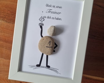 Op de afbeelding: Een wit ingelijst beeld met een zwart-witte tekening van een persoon gemaakt van stenen die een trofee vasthoudt. De tekst in het beeld luidt "Glück ist, einen Trainer wie dich zu haben."