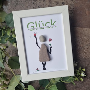 Könnte beinhalten: Gerahmtes Kunstwerk mit weißem Rahmen. Die Kunst zeigt eine Figur aus Steinen, die ein Herz und eine Blume hält. Das deutsche Wort "Glück" steht oben, darunter Text. Grüne Blätter und eine braune Oberfläche im Hintergrund.
