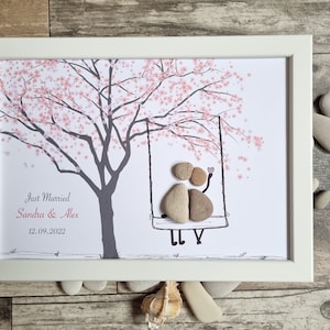 Puede incluir: Impresión artística enmarcada en blanco, que representa un árbol con flores rosas y un columpio con figuras de guijarros. El texto dice "Just Married Sandra & Alex 12.09.2022". El fondo es blanco.