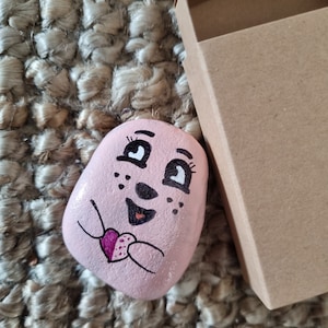 以下が含まれることがあります： ピンクに塗られた石に、漫画の顔、まつげ、そばかすがあります。石はピンクのハートを手に持っています。