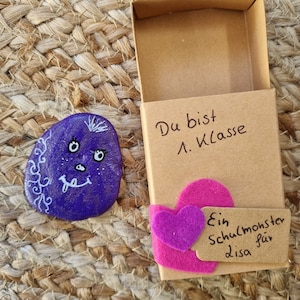 以下が含まれることがあります： 白い顔とピンクのハートが付いた紫色の塗られた石が、茶色の箱の上に置かれています。箱には「Du bist 1. Klasse Ein Schulmonster für Lisa」という文字が書かれています。
