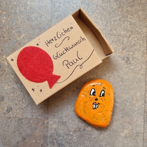 以下が含まれることがあります： 赤いフェルトのハートバルーンと「Herzlichen Glückwunsch Paul」という文字が書かれた小さな茶色の箱。箱の隣には、漫画の顔を描いたオレンジ色の石があります。