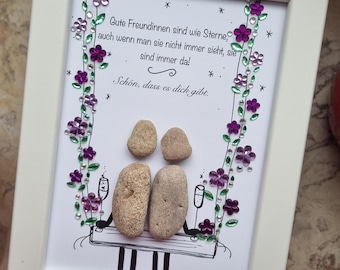 May include: A white framed picture with a hand-painted design of two pebble people holding glasses of champagne on a swing. The background is decorated with purple and green flowers and the text "Gute Freundinnen sind wie Sterne, auch wenn man sie nicht immer sieht, sie sind immer da! Schön, dass es dich gibt."