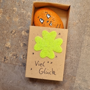 以下が含まれることがあります： 顔と四つ葉のクローバーが描かれた小さな茶色の石が、黒い文字で「Viel Glück」と書かれた茶色の段ボール箱に入っています。