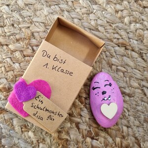 以下が含まれることがあります： 小さな茶色の箱に、ピンクのハートと「Du bist 1. Klasse Ein Schulmonster für Lisa」と書かれた手書きのメッセージがあります。箱が開いており、中には白いハートと顔を描いたピンクに塗られた石が入っています。