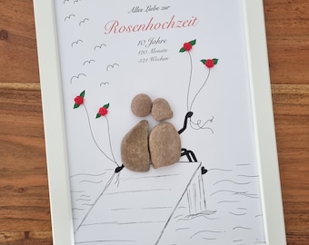 May include: A white framed picture with a black and white drawing of two people sitting on a pier. The drawing is decorated with red and green paper flowers. The text "Alles Liebe zur Rosenhochzeit 10 Jahre 120 Monate 521 Wochen" is printed on the picture.