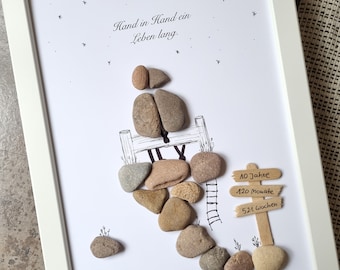 May include: A framed artwork with a hand-drawn illustration of a couple sitting on a bench with a ladder leading up to it. The couple is made of small stones. The artwork has the text "Hand in Hand ein Leben lang." and a sign that reads "10 Jahre, 120 Monate, 521 Wochen."