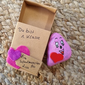 以下が含まれることがあります： 笑顔と赤いハートを描いた小さなピンクの石。石は小さな段ボール箱に入っていて、箱には「Du bist 1. Klasse Ein Schulmonster für Lisa」という手書きのメッセージが書かれています。