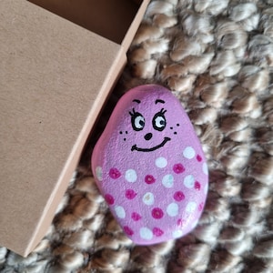 以下が含まれることがあります： 白い水玉模様と笑顔の顔を描いたピンク色の石。石は茶色の織り地の上に置かれています。