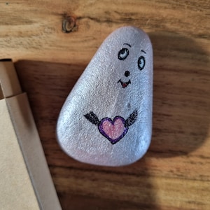 以下が含まれることがあります： 手書きの顔と紫色の羽根のついたピンクのハートが描かれた銀色の石