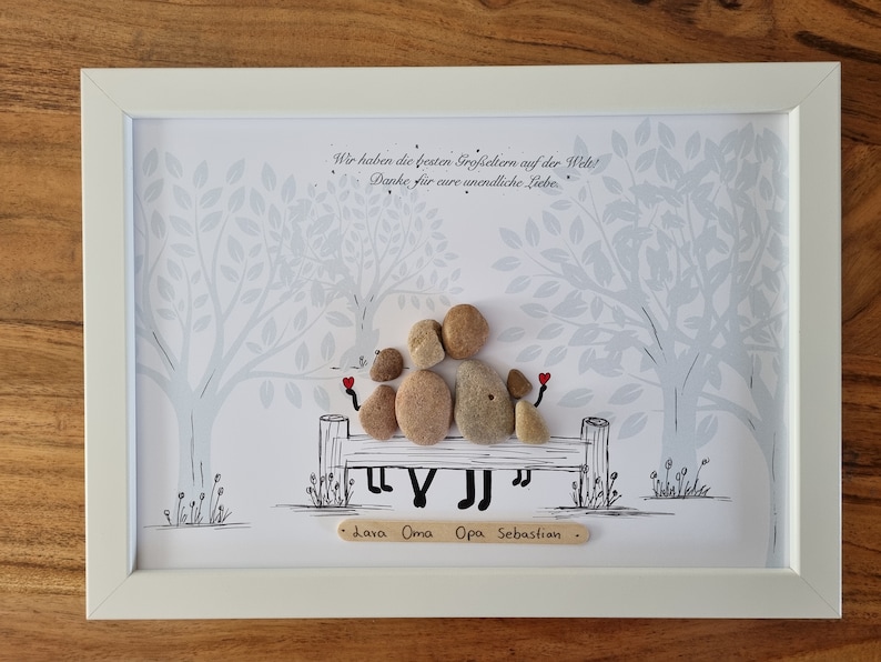 May include: A framed artwork featuring a hand-crafted pebble art design of a family sitting on a bench under a tree. The text "Wir haben die besten Gro&szlig;eltern auf der Welt! Danke f&uuml;r eure unendliche Liebe. Lara Oma Opa Sebastian" is written on the artwork.