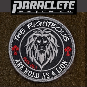 May include: A black and gray embroidered patch with a lion's head in the center. The lion has a white mane and a red cross on each side of its head. The text around the lion reads "The Righteous Are Bold As A Lion" with the scripture reference "Prov 28:1" below the lion. The patch has a gray border.