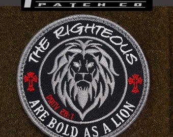 May include: A black and gray embroidered patch with a lion's head in the center. The lion has a white mane and a red cross on each side of its head. The text around the lion reads "The Righteous Are Bold As A Lion" with the scripture reference "Prov 28:1" below the lion. The patch has a gray border.
