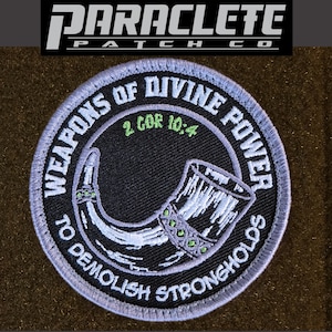 May include: A circular embroidered patch with a black background and a light blue border. It features a white horn illustration, green text reading 2 COR 10:4, and white text reading WEAPONS OF DIVINE POWER TO DEMOLISH STRONGHOLDS around the edge.