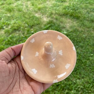 May include: A peach-colored ceramic ring holder with a white ring holder and white cloud designs.