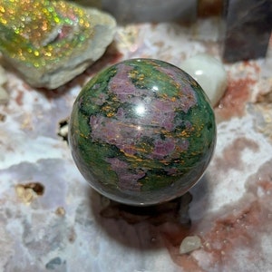 May include: A green, pink, and gold sphere made of polished stone. The sphere is sitting on a white and pink surface.